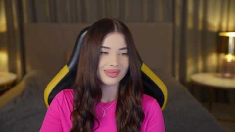 Media: Video of a young woman with long brown hair, wearing a pink shirt, smiling while seated on a black and yellow gaming chair in a dimly lit bedroom with beige curtains and a bedside lamp.