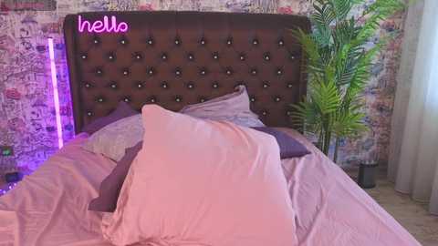 Media: Video of a modern bedroom with a tufted brown headboard, pink pillows, purple bedding, and a vibrant green potted plant. The wall features a mix of brick and abstract art. Neon lights add a pop of color.