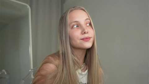 Media: A video of a young, blonde, fair-skinned girl with straight hair, wearing a white top, looking upward thoughtfully against a plain, light-colored background.