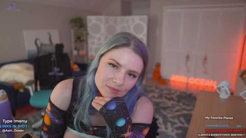 Media: Video of a pale-skinned, blue-haired woman in a black, galaxy-themed outfit, posing seductively on a bed in a dimly-lit room with a desk, orange pumpkins, and a white wardrobe in the background.