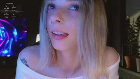 Media: A video of a young, fair-skinned woman with platinum blonde hair, blue eyes, and a septum piercing, wearing a white off-shoulder top, in a dimly-lit room with a TV screen displaying a blue and purple light show in the background.