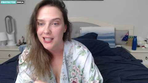 Media: A video of a young woman with long, wavy brown hair and fair skin, wearing a floral robe, sitting on a bed with dark bedding in a modern bedroom.