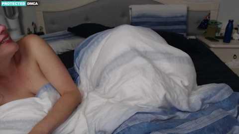 Media: Video of a woman in bed, partially covered by a white and blue striped blanket. Her head is turned to the side, showing a glimpse of her smiling face. The bedroom has a modern, minimalistic design with light-colored walls.