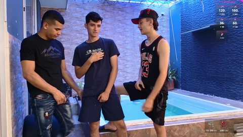 Media: Video of three young men, one in a tank top, two in t-shirts, standing near a small blue swimming pool with a stone wall background.