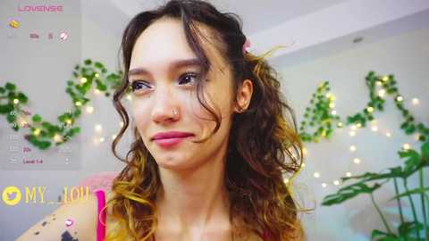 Media: Video of a young woman with curly brown hair, light skin, and pink lipstick, smiling, in a cozy, warmly lit room with string lights, greenery, and \"LOVE\" sign in the background.