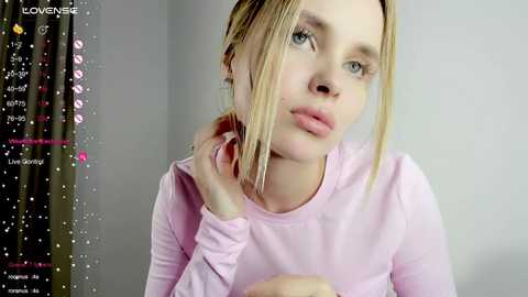 Media: A video of a young Caucasian woman with fair skin, blonde hair, and blue eyes, wearing a light pink long-sleeve shirt. She's in a neutral indoor setting, with a background of muted gray curtains and a digital clock displaying time.