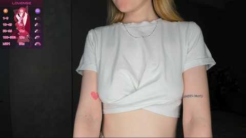 Media: Video of a fair-skinned woman with long blonde hair, wearing a white crop top, showing off a tattoo on her right arm. Background includes a dimly lit room with a purple screen displaying \"LOUNGE.\