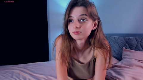 Media: Video of a young Caucasian woman with long brown hair, blue eyes, and fair skin, wearing a beige tank top, lying on a bed with light pink sheets in a dimly lit room.