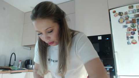 Media: A video of a young woman with long, straight blonde hair, wearing a white t-shirt, cooking in a modern kitchen with white cabinets and a refrigerator adorned with colorful magnets.