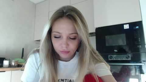 Media: Video of a young Caucasian woman with long blonde hair, wearing a white t-shirt, standing in a modern kitchen with white cabinets and an oven.