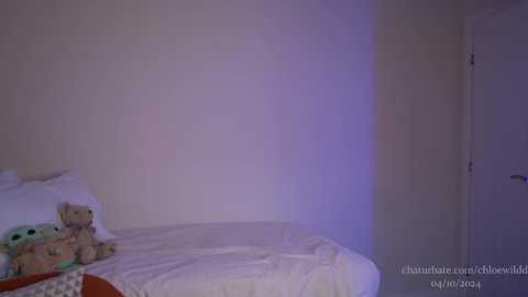 Media: A dimly lit, sparsely furnished bedroom featuring a white bed with a teddy bear and a white pillow, against a plain beige wall. The room is softly lit with purple light.