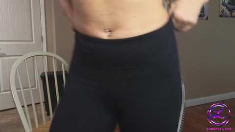 Media: Video of a light-skinned person with a slim waist, wearing black leggings, showing off a navel piercing. Background includes a white door, wooden floor, and a purple circular logo in the bottom right.