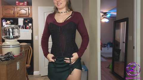 Media: Video of a smiling woman in a burgundy corset top, green mini skirt, and black choker, standing in a kitchen with wooden cabinets, stove, and a mirror.