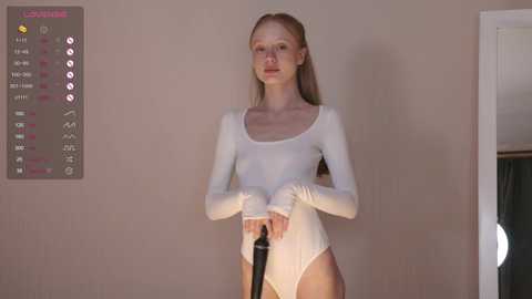 Media: Video of a slender, fair-skinned, blonde woman in a white, long-sleeved leotard, holding a black hairbrush, against a beige wall with a digital display showing \"Lipstick,\" \"Eye shadow,\" and \"Blush\" in pink.