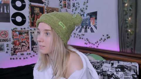 Media: Video of a blonde woman with fair skin wearing a green beanie, off-shoulder white shirt, and hanging out in a colorful, cluttered bedroom with posters, records, and fairy lights.