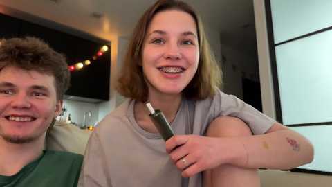 Media: Video of a smiling young woman with braces, light skin, and shoulder-length brown hair, holding a green lighter in a cozy, modern kitchen.