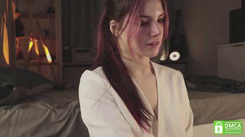 Media: Video of a young Caucasian woman with long, straight, dark red hair in a ponytail, wearing a white robe, standing in a dimly lit bedroom with a bed, nightstand, and lamp.