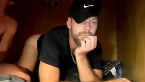 Media: Video of a muscular man with a beard and black cap, leaning on his elbow, in a dimly lit room.