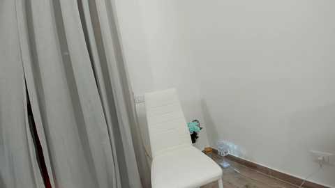 Media: Video of a minimalist, sparsely furnished room with a white cushioned chair against a plain white wall. Gray curtains cover a window on the left, and a plastic bag is on the floor near the chair.