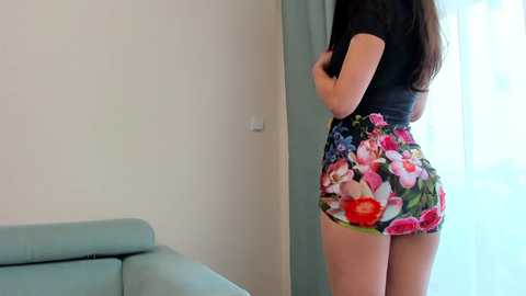 Media: Video of a woman with long brown hair, wearing a black top and floral-patterned shorts, standing beside a light green sofa in a minimalist room with white walls and sheer curtains.