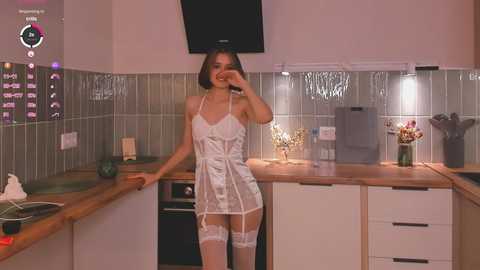 Media: Video of a young woman with a slim physique, wearing a sheer white lace lingerie set with matching garter belt, posing seductively in a modern kitchen.