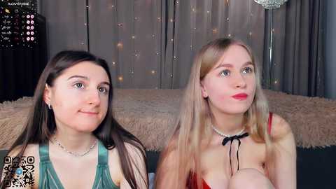 Media: Video of two young women with fair skin, long straight hair, and minimal makeup, seated on a beige couch. One wears a green top, the other a red lace dress, both with jewelry. Background features a bed with a fur throw and a glittery chandelier.