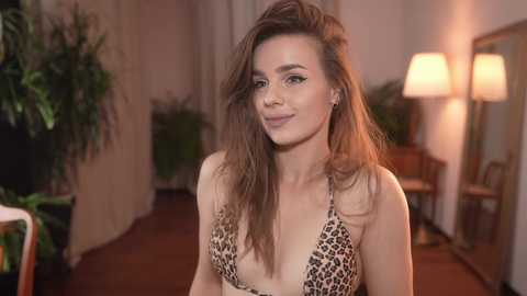 Media: Video of a smiling Caucasian woman with long brown hair, wearing a leopard-print bikini top, standing in a warmly lit, indoor setting with potted plants and a lit floor lamp in the background.