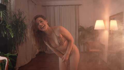 Media: Video of a smiling woman in leopard-print bikini, leaning forward, in a dimly lit room with a potted plant, wall lamps, and curtains.