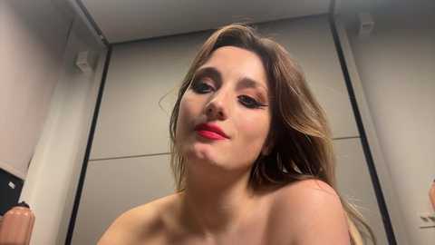 Media: Video of a fair-skinned woman with long, wavy brown hair, wearing bright red lipstick and eye makeup, topless, in a dimly lit room with gray walls.