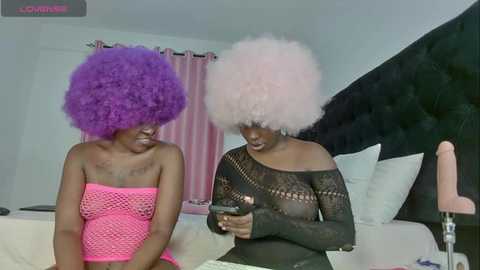 Media: Video of two Black women with voluminous, exaggerated hair in bright purple and pink, wearing revealing, patterned outfits in a modern bedroom with white walls and a dark headboard.