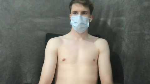 Media: Video of a young, fair-skinned man with short brown hair, wearing a light blue surgical mask, seated against a dark, textured wall.