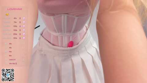 Media: A close-up video of a woman wearing a pink corset over a white pleated skirt, with a QR code in the lower left corner. The image captures her torso and waist, emphasizing the corset's intricate detailing.