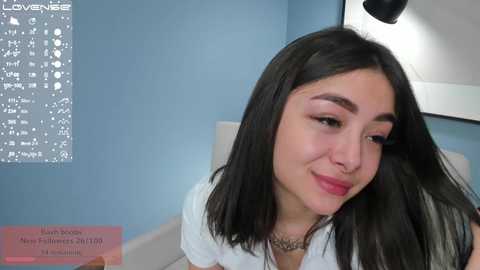 Media: A video of a smiling young woman with long black hair, light skin, and dark eyes, wearing a white blouse, in a modern, blue-walled room.