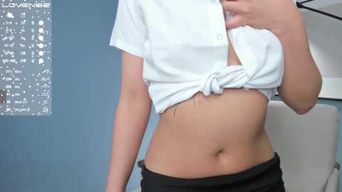 Media: A video of a woman in a white, short-sleeved top tied in a knot, revealing her midriff and black pants. Background includes a light blue wall with white \"Love\" text and a modern chair.