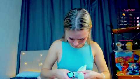 Media: Video of a young woman with blonde hair, wearing a light blue sleeveless top, playing a video game on a bed with a white headboard. The background features a dark curtain and a colorful toy shelf.