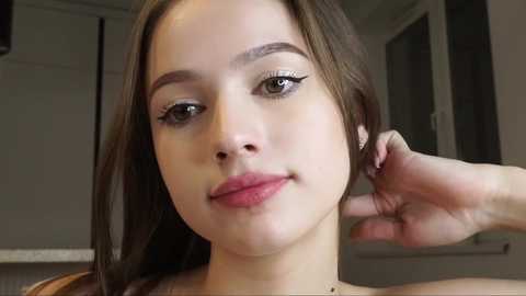 Media: Video of a young woman with fair skin, straight brown hair, and light makeup, wearing a brown top, touching her ear. The background shows a modern kitchen with white cabinets and a window.