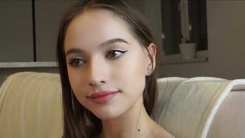 Media: Video of a young, fair-skinned woman with straight brown hair, wearing subtle makeup, and sitting on a beige cushioned chair. The background shows a modern kitchen with gray cabinets and a small white plant.