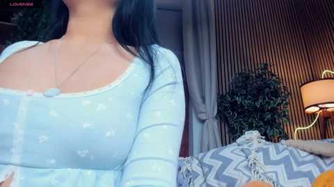 Media: Video of a woman with long black hair, wearing a light blue long-sleeved shirt with a heart pattern, standing near a bed with a blue and white zigzag blanket, a potted plant, and a lamp.