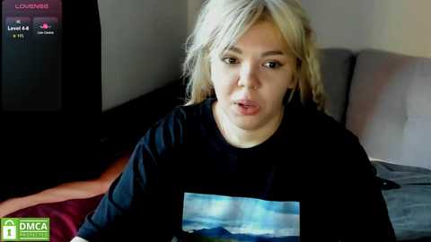 Media: Video of a fair-skinned woman with blonde hair, wearing a black T-shirt with a graphic, seated on a grey couch, watching a TV screen displaying a news ticker.