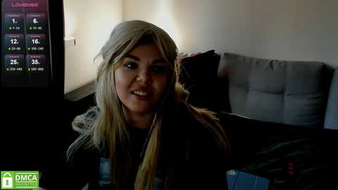 Media: Video of a smiling young woman with long blonde hair, wearing a dark jacket, sitting in a dimly-lit room with a gray couch and TV displaying \"LOVEMIX\" and \"DICA\" logos.