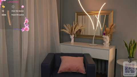 Media: Video of a modern living room with a dark blue sofa, a white console table with a gold-framed mirror, and decorative white and gold vases. Soft pastel-colored curtains and a pink throw pillow add a cozy touch.