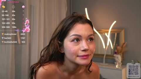 Media: Video of a young woman with long brown hair, fair skin, and a soft smile, sitting in a modern room with a white vase and a mirrored wall, a QR code visible on the right.