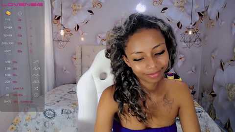 Media: A video of a young woman with medium brown skin and curly hair, wearing a purple strapless top, sitting in a modern room with floral wallpaper and hanging lights.