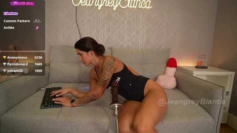 Media: Video of a tattooed woman in a black bodysuit, lounging on a grey sofa, typing on a laptop, with a red dildo on the couch.