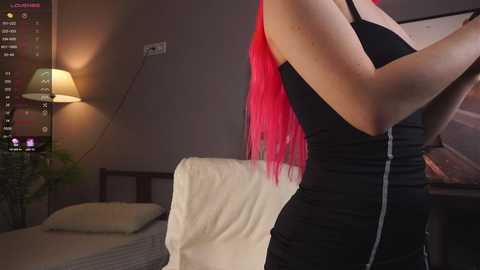 Media: Video of a fair-skinned woman with long, vibrant pink hair, wearing a black sleeveless dress, standing in a dimly lit bedroom with a white bed, a lamp, and a potted plant.