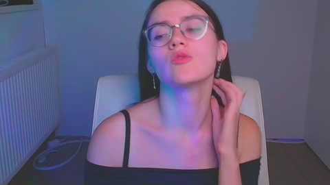 Media: Video of a young woman with fair skin, straight black hair, and large glasses, wearing a black off-shoulder top, kissing her hand, seated in a white chair against a blue and purple-lit background.