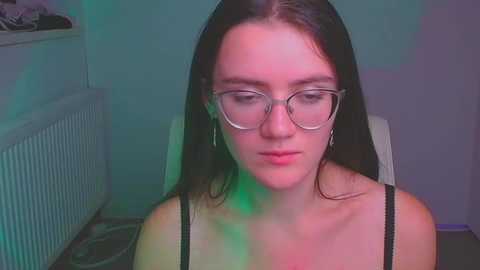 Media: Video of a young woman with fair skin and long dark hair, wearing glasses and a black top, sitting in a chair with a green and purple light shining on her face.