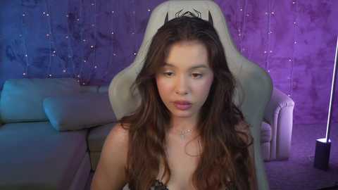 Media: Video of a young woman with long, wavy brown hair, wearing a leopard-print bra, seated in a beige gaming chair in a dimly lit room with purple and blue lighting, textured walls, and a couch.