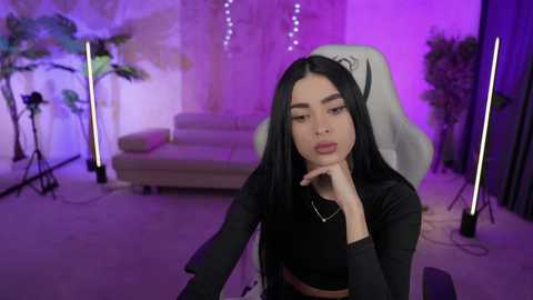 Media: Video of a young Asian woman with long black hair, wearing a black crop top, sitting on a gaming chair in a modern, purple-lit room with plants and studio lights.