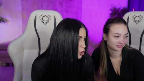 Media: Video of two women, one with long black hair, the other with light brown hair, sitting side by side in white gaming chairs with black skull logos, under purple lighting.